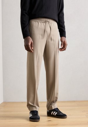 Pantalones beige, ajustados con cintura elástica y cordón. Material suave, corte holgado y piernas rectas. Combinados con zapatillas negras.