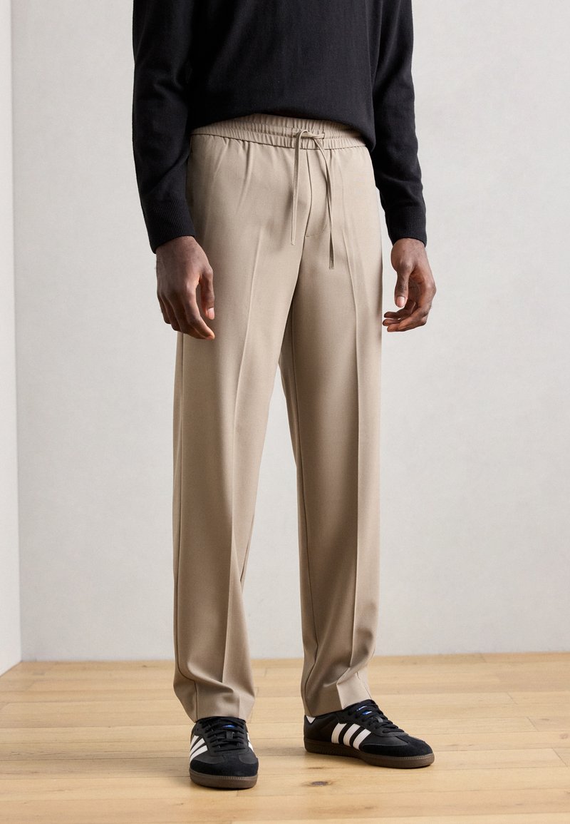 Pantalons beiges ajustés avec une taille élastique et un cordon de serrage. Matériau lisse, coupe ample et jambes droites. Portés avec des baskets noires.