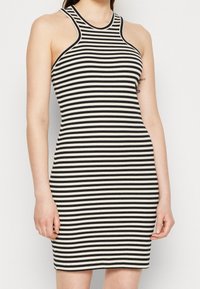 Černobílé pruhované šaty s vysokým výstřihem, bez rukávů a s přiléhavým střihem. Žebrovaná textura zvyšuje vizuální zajímavost.