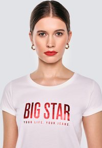 Mladá žena s hladce učesanými vlasy, červenou rtěnkou a zlatými náušnicemi ve tvaru kruhu, oblečená v bílé košili s červeným potiskem "BIG STAR Váš život. Vaše džíny."