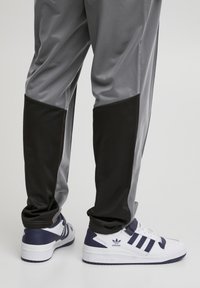 Pantalones deportivos grises con acentos negros y una textura suave, combinados con zapatillas blancas y marinas que tienen tres franjas y un logo.