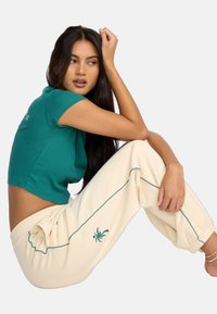 Teal, krótka bluzka z krótkim rękawem, zestawiona z kremowymi spodniami dresowymi z turkusowymi lamówkami i dekoracyjnym motywem palmowym.