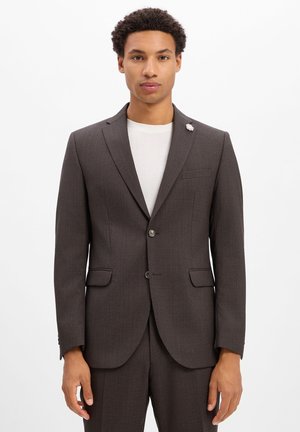Mann mit kurzen lockigen Haaren, der eine dunkelbraune Anzugjacke über einem weißen Hemd trägt und vor einem einfarbigen weißen Hintergrund steht.