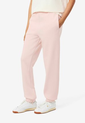 Personne portant un pantalon de survêtement rose clair et des baskets blanches, debout avec une main dans la poche sur un fond blanc uni.