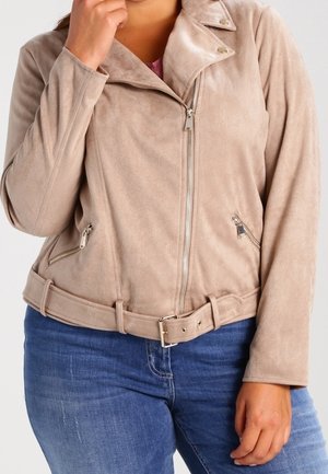 Vrouw draagt een beige suède jas met ritssluiting, voorzien van een riem en twee ritssluitzakken, gecombineerd met een blauwe spijkerbroek.