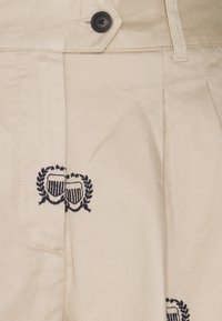 Béžové bavlněné kalhoty s černým knoflíkem, sklady na přední straně a vyšitým erbem s vavřínovými akcenty u kapsy.