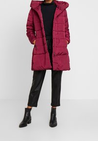 Burgunderfarbener Puffer-Mantel mit Kapuze, mit einem gesteppten Design und Frontreißverschluss. Kombiniert mit schwarzen Lederhosen und Ankle-Boots.