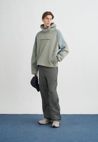 Υπερμεγέθης πράσινη κουκούλα με μπλε λεπτομέρειες, γκρί cargo παντελόνι και ανοιχτό γκρί παπούτσια. Η κουκούλα διαθέτει ψηλό λαιμό και λογότυπο. Κρατάει ένα σκούρο καπέλο.