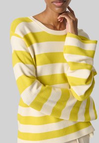 Pull rayé en jaune et crème, texture côtelée, encolure dégagée et coupe ample avec des épaules tombantes. Contraste de couleurs notable dans les rayures.