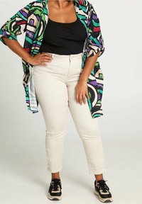 Chemise oversize multicolore à motifs abstraits avec du vert, du violet et du noir, portée sur un débardeur noir. Pantalon beige ajusté et baskets noires.