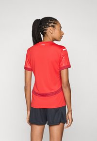 Red athletic jersey with short, textured sleeves and a solid back. Features a round neckline and a contrasting maroon accent at the hem.