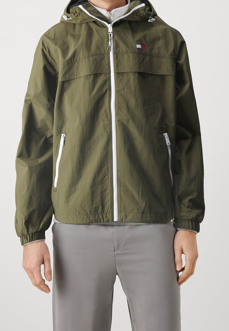 Olivgrön lätt jacka med dragkedja fram, elastiska ärmslut och huva. Har vit dragkedja och logotyp på bröstet. Slät textur.