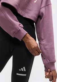 Κοντή, φαρδιά μπλούζα φούτερ σε ανοιχτό μωβ χρώμα, με ριμπ μανσέτες και γραφικό σχέδιο. Συνδυάζεται με μαύρο κολάν ψηλόμεσο με λεπτομέρεια λογοτύπου.