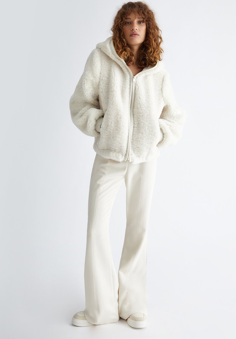 Femme aux cheveux bouclés portant une veste en peluche blanche zippée, un pantalon large crème et des baskets blanches sur un fond léger uni.