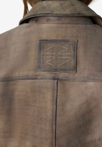 Vue arrière d'une veste en daim marron avec un patch carré cousu arborant un logo géométrique embossé centré sous le col.