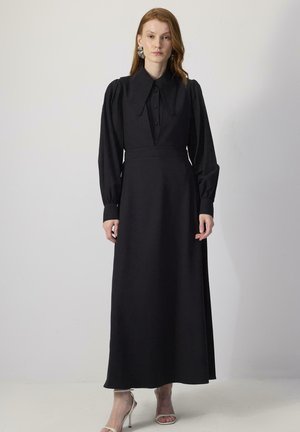 Femme aux cheveux roux portant une longue robe noire avec un grand col, des manches bouffantes et des sandales blanches à talons hauts, debout contre un mur uni.