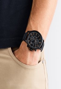 Mustari musta kello tekstureeritud musta kelladialiga, oranžide aktsentidega ja mustast silikoonist rihmaga, kantud randmel khaki kangast backgroundil.