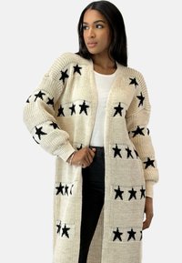 Cárdigan de punto crema con patrones de estrellas negras, de frente abierto, mangas largas y puños acanalados. Textura suave, corte holgado y diseño de longitud media.