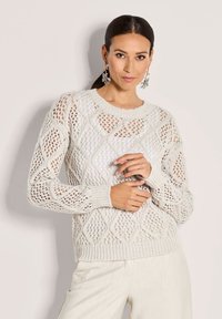 Eine Frau mit dunklem Haar trägt einen weißen, offen gestrickten Pullover mit Diamantmustern, kombiniert mit hellen Hosen und hängenden Ohrringen in Form von Blättern.