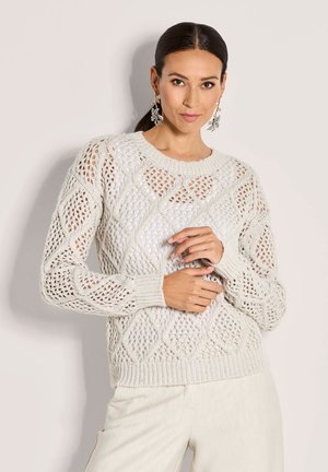 Eine Frau mit dunklem Haar trägt einen weißen, offen gestrickten Pullover mit Diamantmustern, kombiniert mit hellen Hosen und hängenden Ohrringen in Form von Blättern.