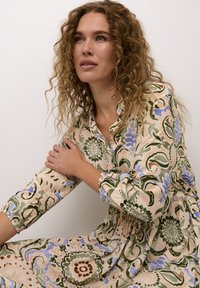 Blommönstrad klänning i beige, med gröna och blå mönster. Långa ärmar med elastiska manschetter och detalj med midjeband. Mjuk tygkvalitet.