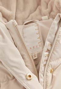 Manteau en duvet beige avec doublure en peluche, fermeture éclair à l'avant et quincaillerie dorée. Présente une étiquette avec "BAKER" en doré, spécifications d'âge sur l'étiquette.