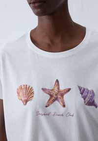 Vit bomulls t-shirt med ett grafiskt motiv av tre snäckor: en kammussla, sjöstjärna och spiralformad snäcka, med texten "Seashell Beach Club" nedanför.
