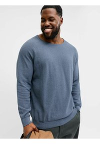 Blauer, strukturierter Pullover mit weitem Ausschnitt und langen Ärmeln, mit einem gerippten Saum versehen. Das Model hält einen beigen Gegenstand, vor einem hellen Hintergrund.