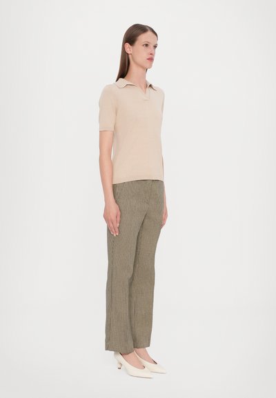 Polo beige a maniche corte con colletto abbinato a pantaloni a vita alta a quadri verdi e tacchi bianchi, in piedi su uno sfondo bianco.