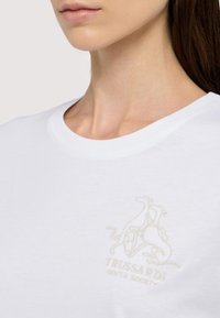 Camiseta de algodón blanca con un sutil logotipo bordado en beis de dos galgos y el texto "TRUSSARDI GENTLE SOCIETY" debajo. Manga corta, cuello redondo.