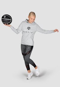 Felpa grigia con cappuccio e logo, abbinata a leggins neri e grigi con accenti in rete, e scarpe da ginnastica bianche. Tiene un pallone da basket.