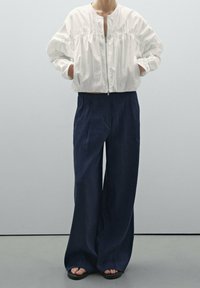 Une personne portant une veste blanche zippée avec les mains dans les poches, un pantalon large bleu marine et des sandales noires, se tenant contre un fond blanc.