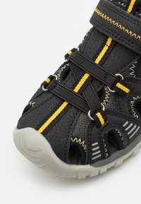 Musta urheilukenki, jossa on keltaisia yksityiskohtia, verkkopaneelit, säädettävät hihnat ja kuvioitu kumipohja. Kiinnitys joustavilla nauhoilla.