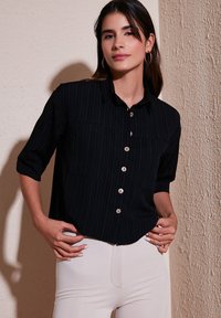 Black striped button-up shirt with two front pockets, wooden buttons, and three-quarter sleeves, paired with light-colored pants.