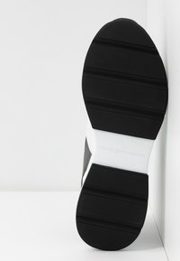 Suola di scarpa in gomma nera con scanalature testurizzate e un accento bianco. Il materiale sembra resistente con un design piatto per la stabilità.