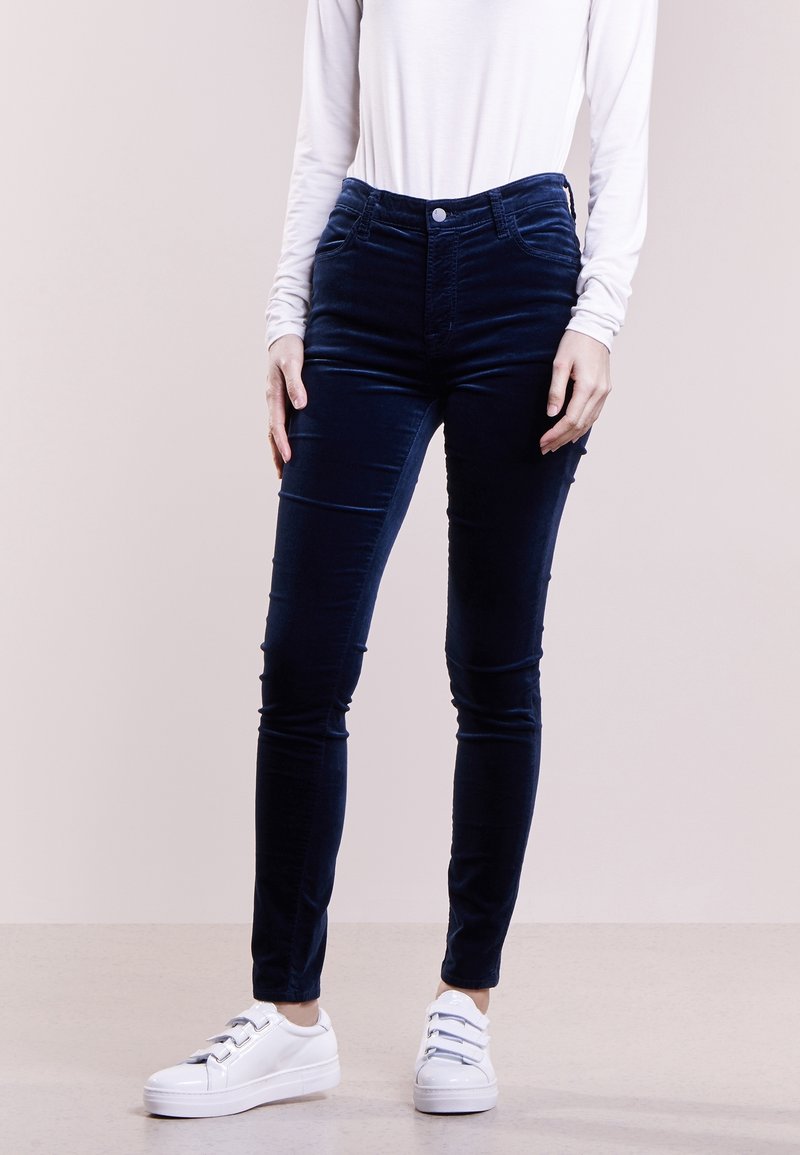 Personne portant un pantalon bleu foncé ajusté, un haut blanc à manches longues et des baskets blanches à scratch debout sur un sol clair.