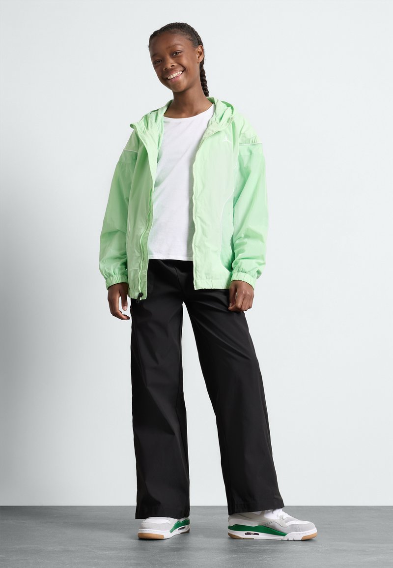 Giacca a vento verde chiaro con polsini elastici, t-shirt bianca, pantaloni neri a gamba larga e scarpe bianche con dettagli verdi.