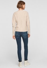 Beige gebreide trui met een losse pasvorm, gecombineerd met donkerblauwe skinny jeans en crèmekleurige sneakers, gezien vanuit de achterkant.