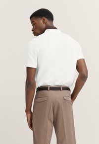 Weißes Poloshirt mit kurzen Ärmeln und gerippter Struktur, kombiniert mit braunen, mit einem Gürtel versehenen Hosen mit zwei aufgesetzten Gesäßtaschen.