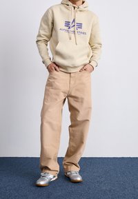 Sudadera con capucha beige con logo púrpura, combinada con pantalones holgados beige claro y zapatillas plateadas. Textura de tela suave y ajuste relajado.