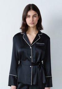 Czarna satynowa bluzka piżamowa z białym wykończeniem, długie rękawy, kołnierzyk, kieszeń z przodu oraz przewiązywany pas. Zapięcie na guziki z przodu.