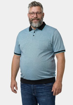 Hymyilevä keski-ikäinen mies, jolla on harmaa parta ja silmälasit, pukeutunut vaaleansiniseen poolopaitaan, jossa on musta kaulus ja reunukset, sekä sinisiin farkkuihin.