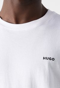 Wit katoenen T-shirt met ronde hals en een zwart "HUGO"-logo gedrukt op de linkerborst. Gladde textuur, standaard pasvorm.