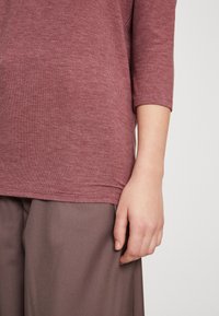 Top in misto cotone di colore rosa con maniche a 3/4. Il tessuto ha una consistenza morbida e un motivo in maglia sottile e uniforme. Mano parzialmente visibile.