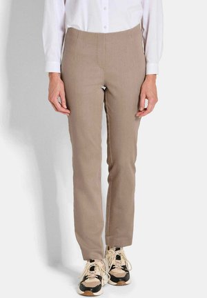 Vrouw in beige strakke broek, witte blouse met lange mouwen en beige en zwarte sportschoenen, staand tegen een effen achtergrond.
