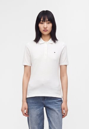 Jeune personne aux cheveux noirs et raides portant un polo blanc à manches courtes et un jean bleu sur un fond uni.