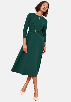 Grünes, knielanges Kleid mit langen Ärmeln und einem Schlüsselloch-Ausschnitt. Es verfügt über ein verdrehtes Taillendetail und eine goldene Schnalle als Akzent, hergestellt aus glattem Stoff.