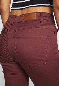 Vaqueros skinny burdeos con una textura suave, que presentan bolsillos traseros, un parche de cuero marrón en la cintura y sutiles detalles de costura.