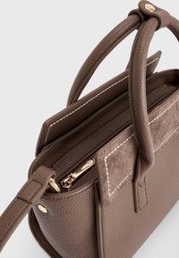 Bolso de cuero marrón texturizado con asas dobles, costuras blancas, acento de ante y tirador dorado en la cremallera, fotografiado sobre un fondo blanco.
