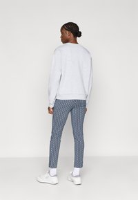 Pelēka sporta jaka ar brīvu piegriezumu, zili bikses ar ģeometrisku rakstu, un baltas sporta apavus. Modelis redzams no aizmugures.